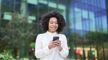 Genç Afro-Amerikan bayan çalışanı ofis binasının yakınındaki sokakta yürürken akıllı telefon kullanıyor. Gülümseyen siyahi kadın mesaj yazıyor, kaydırıyor, sohbet ediyor ya da sosyal medyada geziniyor