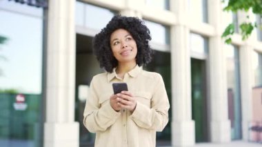 Genç Afrikalı Amerikalı kız öğrenci üniversite binası yakınlarındaki kampüste yürürken akıllı telefon kullanıyor. Gülümseyen siyahi kadın mesaj yazıyor, internette sohbet ediyor ya da sosyal medyada geziniyor
