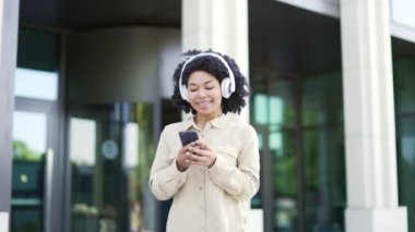 Mutlu Afro-Amerikalı kız öğrenci üniversite binasının yakınındaki kampüste yürürken kulaklıkla müzik dinliyor. Gülümseyen siyah kadın çalma listesini açıyor ve akıllı telefondan şarkı söylemekten hoşlanıyor.