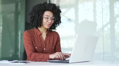 Genç Afro-Amerikan bayan çalışanı iş yerindeki iş yerinde dizüstü bilgisayarında oturuyor. Gözlüklü, gülümseyen siyah bir kadın bir projede bilgisayarda çalışıyor, internette sohbet ediyor, e-posta yazıyor.