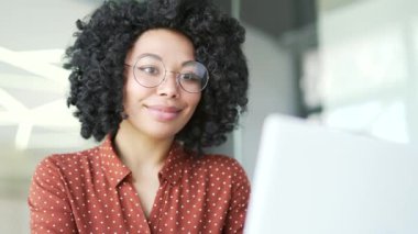 Kapatın. Genç Afro-Amerikan bayan çalışanı iş yerindeki iş yerinde dizüstü bilgisayarında oturuyor. Gözlüklü siyahi kadın bir projede bilgisayarda çalışıyor, internette sohbet ediyor, e-posta yazıyor.