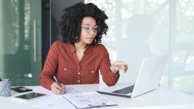 Genç Afro-Amerikan kadın muhasebeci ya da finansör ofiste bir kalemle dokümanları dolduruyor. İş kadını dizüstü bilgisayardaki verileri kontrol ediyor. Siyah kadın boş bir vergi formu dolduruyor.