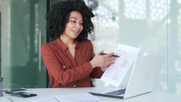 自信を持ったアフリカ系アメリカ人女性マネージャーは オフィスの職場に座っているノートパソコンを使ってビデオ通話でオンラインで話します リモートプレゼンテーションの財務報告書と統計資料を示しています — ストック動画