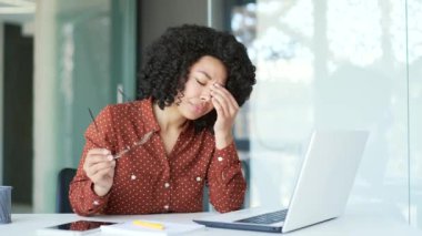 İş yerinde dizüstü bilgisayarla çalışırken göz yorgunluğu ve baş ağrısından muzdarip çok çalışan Afrikalı Amerikalı genç bayan çalışanlar. Yorgun siyah kadın gözlüğünü ve masajını çıkarıyor, burun köprüsünü ovuyor.