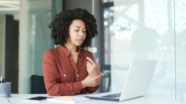 Bilek ağrısından muzdarip genç Afro-Amerikan bayan çalışanı iş yerinde masa başında dizüstü bilgisayarla çalışıyor. Zenci bir kadın esrara masaj yapıyor, elleriyle egzersiz yapıyor.