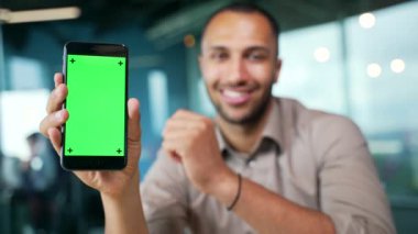 Modern ofiste oturan genç iş adamını gösteren yeşil ekranlı akıllı telefonu kapat. Renk anahtarı, reklam için dikey şablon düzeni. Gülümseyen karışık ırk adamı telefonu işaret ediyor.