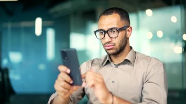 Ciddi odaklı gözlüklü bir işadamı cep telefonu kullanıyor modern ofiste masa başında oturuyor. Yakışıklı İşveren Yöneticisi, müşterisiyle uzaktan sörf sorunları çözer, yeni iş uygulaması öğrenir. Kapat