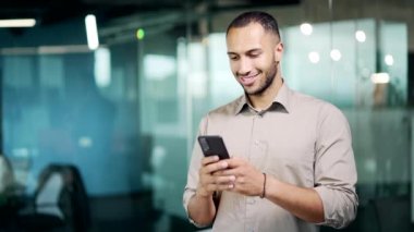 Yakışıklı iş adamı el ele tutuşan bir cep telefonu kullanıyor. Mutlu erkek serbest çalışan, sohbet eden, daktilo kullanan modern cam ofis çalışma yerinde duran akıllı telefon kullanan. Gülümse.