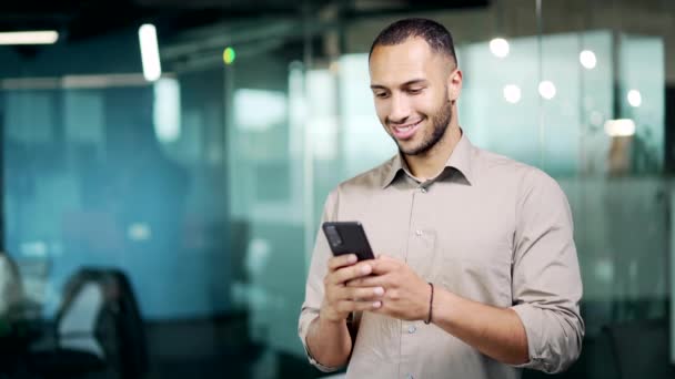 英俊的商人手里拿着手机 快乐的男性自由职业者员工男人站在现代玻璃办公室室内用智能手机浏览聊天发短信打字 — 图库视频影像