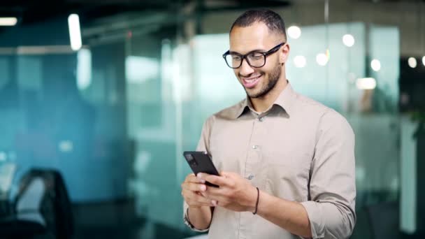 英俊的商人手牵着手 用手机 快乐的男性经理员工站在现代玻璃办公室的室内 戴着眼镜 用智能手机浏览聊天短信 — 图库视频影像