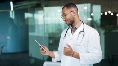 Tıp önlüklü neşeli bir doktor video görüşmesi için tablet bilgisayar kullanıyor ve klinikte çalışan gözlüklü bir doktor olan hasta ile çevrimiçi istişare yapıyor. terapi.