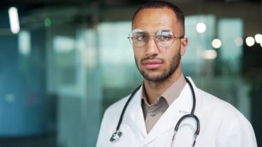 Modern ofis kliniğinde dikilirken kameraya bakan kendine güvenen genç bir doktor portresi. Gözlüklü ve beyaz önlüklü yakışıklı melez doktor.