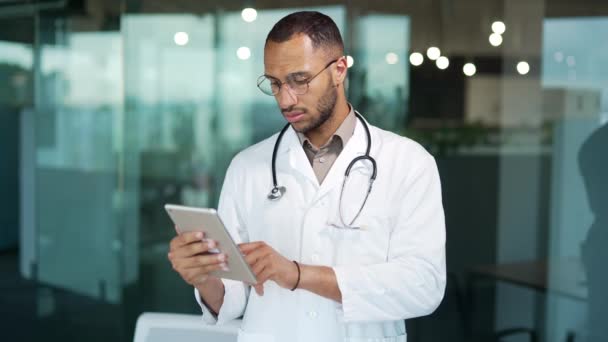 形象严肃的医生使用数码平板站在现代诊所的办公室 戴眼镜 白衣打字 浏览聊天 阅读电子邮件等 自信地专注于混合种族的医生 — 图库视频影像