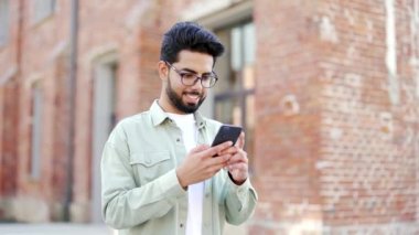Yakışıklı sakallı bir öğrenci üniversite binasının yanındaki kampüs alanında dururken akıllı telefon kullanıyor. Mutlu gülümseyen erkek sohbeti, sosyal medyada gezinme, mesaj atma veya okuma