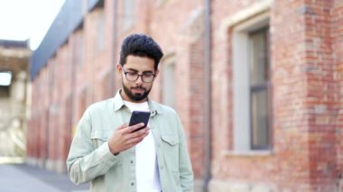 Yakışıklı sakallı bir öğrenci üniversite binasının yanında kampüs çevresinde yürürken akıllı telefon kullanır. Mutlu gülümseyen erkek sohbeti, sosyal medyada gezinme, mesaj atma veya okuma
