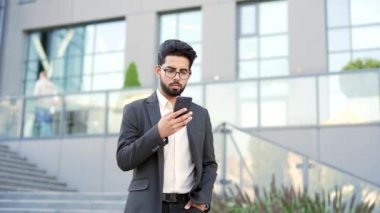 Resmi takım elbiseli gülümseyen genç işadamı bir ofis binasının yakınında yürürken akıllı telefon kullanıyor. Mutlu girişimciler telefonlarını karıştırıyor, e-posta mesajlarını okuyor ya da internette sohbet ediyor.