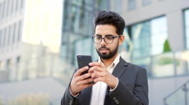 Resmi takım elbiseli gülümseyen genç işadamı, bir ofis binasının yanındaki caddede duran akıllı telefonu kullanıyor. Mutlu girişimci telefonuna bakıyor, e-posta mesajlarını kontrol ediyor ya da internette sohbet ediyor. Kapat.
