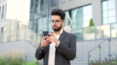 Resmi takım elbiseli mutlu genç bir işadamının portresi caddede bir ofis binasının yanında duruyor. Elinde akıllı telefonla kameraya bakarak poz veren gülümseyen bir girişimci. Müdürün kafasından.