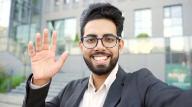 Bakış açısı. Resmi takım elbiseli gülümseyen genç bir işadamı, bir video görüşmesinde bir ofis binasının yakınında duran akıllı telefondan selfie çekiyor. Kameraya bakarak iletişim kuran mutlu bir erkek.