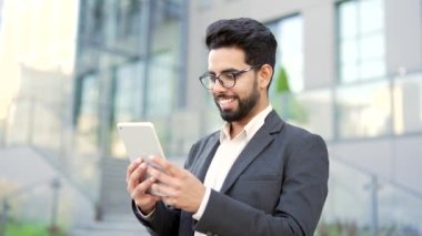 Resmi takım elbiseli gülümseyen genç işadamı, bir ofis binasının yanında sokakta dururken tablet kullanıyor. Mutlu girişimciler e-postaları kontrol eder, sohbet eder, mesajlaşır veya internetten alışveriş yapar.