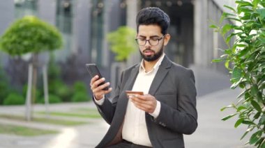 Şok olmuş genç işadamı, ofis binasının yanındaki bankta oturan akıllı telefondan kredi kartını kontrol ediyor. Hüsrana uğramış işçi dolandırıcılıkla yüzleşti, hesabından para çalındı. O bir aldatma kurbanı oldu.