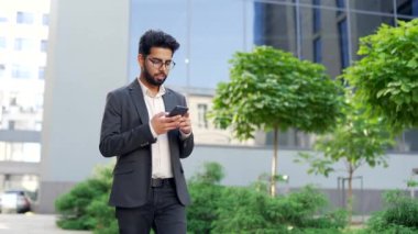 Resmi takım elbiseli gülümseyen genç işadamı bir ofis binasının yakınında yürürken akıllı telefon kullanıyor. Mutlu girişimciler telefonlarını karıştırıyor, e-posta mesajlarını okuyor ya da internette sohbet ediyor.