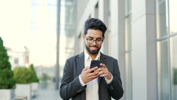 笑着穿正装的年轻商人在办公楼附近的街上散步时 用的是智能手机 兴高采烈的创业者在网上浏览电话 阅读邮件或聊天 — 图库视频影像