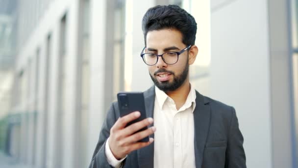 身穿正装的年轻商人站在一幢办公楼附近的街上 一边笑一边用智能手机 兴高采烈的创业者在网上浏览电话 阅读邮件或聊天 — 图库视频影像