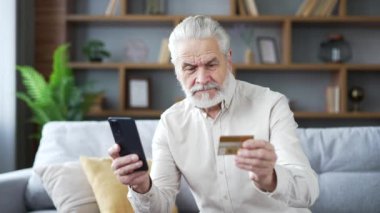 Mutlu yaşlı bir adam online alışveriş yapıyor. Evdeki kanepede oturan akıllı telefondan kredi kartı numarasını yazıyor. Gülümseyen yetişkin erkek indirimlerden, terfilerden, satışlardan tatmin olan bir alışveriş yapar.