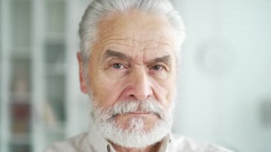 Evdeki oturma odasında duran ciddi, gri saçlı, yaşlı bir adamın portresini yakından çek. Yakışıklı emekli erkek kameraya bakarak poz veriyor. Özgüvenli, düşünceli, sakallı emeklinin vesikalığı.