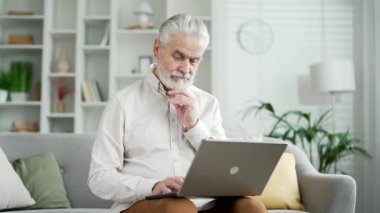 Evdeki oturma odasındaki kanepede dizüstü bilgisayarda çalışan çok düşünceli yaşlı bir işadamı. Olgun erkek mesajlaşması, internette gezinme, internette sohbet bankacılığı uygulaması.
