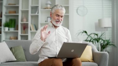 Evde oturma odasındaki kanepede otururken bilgisayarla video görüşmesinde konuşan yakışıklı, yaşlı, kır saçlı adam. Olgun gülümseyen yetişkin erkek çevrimiçi sohbet ediyor, uzaktan görüşme.