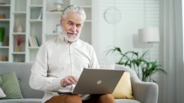 Mutlu, gri saçlı yaşlı adam evdeki oturma odasındaki kanepede oturan dizüstü bilgisayarından harika haberler aldı. Gülümseyen yetişkin erkek bilgisayarda hoş bir mesaj okuyup başarıyı kutluyor.