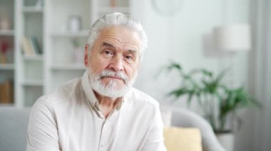 Evdeki parlak bir odada, koltukta oturan gri saçlı, ciddi bir yaşlı adamın portresi. Yakışıklı emekli erkek kameraya bakıyor. Kendinden emin gülümseyen olgun sakallı büyükbabanın fotoğrafı.