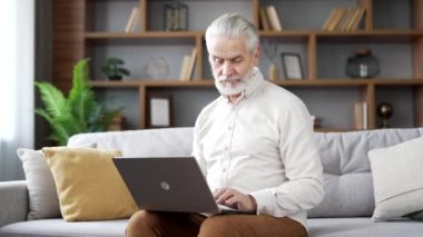 Dizüstü bilgisayarda çalışan yaşlı bir adam evdeki oturma odasında kanepede oturuyor. Olgun iş adamı mesajlaşması bilgisayardan mesajlaşma internette gezinme, online alışveriş, sohbet, uygulama bankacılığı