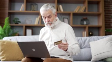 Mutlu yaşlı adam online alışveriş yapıyor. Evdeki kanepede oturan dizüstü bilgisayarında kredi kartı numarasını yazıyor. Gülümseyen yetişkin erkek indirimlerden ve terfilerden memnun kalarak alışveriş yapıyor.