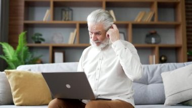 Kafası karışmış gri saçlı yaşlı adam evdeki oturma odasındaki kanepede otururken dizüstü bilgisayar kullanmakta zorlanıyor. Kafası karışmış yetişkin erkeğin bilgisayardaki uygulamayla ilgili karmaşık sorunları var.