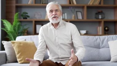 Webcam görüntüsü. Memnun olmayan yaşlı, yaşlı, gri sakallı adam evdeki oturma odasındaki kanepede oturmuş video görüşmesinde konuşuyor. Kızgın olgun erkek kameraya bakmaktan şikayetçi.