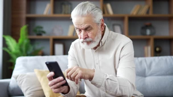 自宅のリビングルームにソファーに座っているスマートフォンを使用して困難な灰色の髪を持つ混乱した老人 心配した成熟した男性は 電話でのアプリケーションに取り組む問題の複雑さを持っています — ストック動画