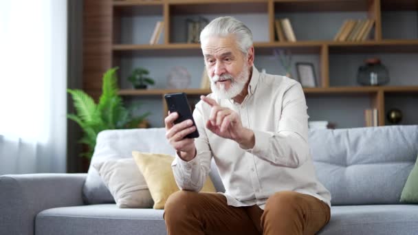 灰色の髪を持つ幸せな興奮する高齢男性は 自宅のリビングルームにソファーに座っている間 スマートフォンで素晴らしいニュースを受け取りました 大人の成熟した男性は電話で楽しいメッセージを読み 成功を祝います — ストック動画