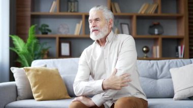 Oturma odasındaki kanepede oturan dirsek ağrısından muzdarip yaşlı gri saçlı adam. Üzgün, emekli bir erkek bir esrara masaj yapıyor, elini ovuyor. Eklem yaralanması ya da bağ burkulması var.