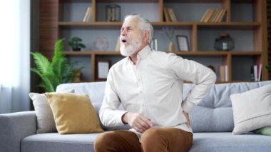 Yaşlı, yaşlı, kır saçlı, sırt ağrısı çeken adam evdeki oturma odasında kanepede oturuyor. Olgun emekli erkek masajı, ağrılı bel altı kasları. Yorgun ya da siyatiği var.