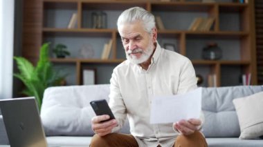 Mutlu kıdemli adam evde otururken akıllı telefon kullanarak faturaları ödemek için mobil uygulama kullanıyor. Olgun erkek, ev ekonomisini hesaplar ve verileri sohbet robotuna aktarır. Ödemesi kolay ve basit.