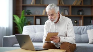 Yaşlı, yaşlı, kır sakallı adam evdeki oturma odasında kanepede otururken internetten e-öğrenme, video kursu dinliyor. Yetişkin bir erkek uzak mesafeden öğrenir ve not defterine yazar.