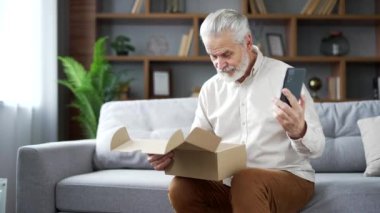 Yaşlı kıdemli bir adam evde hediye paketi alıyor ve akıllı telefondan internet üzerinden konuşuyor. Gülümseyen neşeli erkek çevrimiçi bir mağazadan alışveriş yapmaktan mutlu. E-ticaretin tatmin olmuş müşterisi