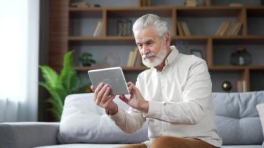 Yaşlı yaşlı yaşlı gri adam evdeki oturma odasında kanepede otururken dijital tablet kullanarak göz gezdiriyor. Yetişkin, emekli bir erkek video akışı, çevrimiçi yayın veya sosyal ağlar izliyor