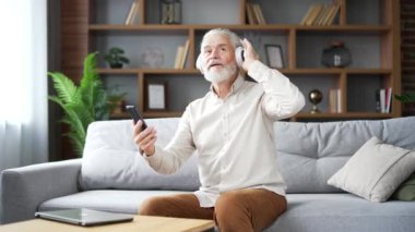 Kablosuz kulaklıklı yaşlı gri sakallı adam evdeki oturma odasında kanepede oturmuş müzik dinliyor. Tişört giymiş olgun bir erkek, akıllı telefonunda çalma listesini açar ve şarkının tadını çıkarır.