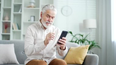 Evde oturma odasındaki kanepede oturan gri saçlı, akıllı telefonlu yaşlı bir adam. Yetişkin emekliler okur, mesajlar yazar, sohbet eder, online alışveriş yapar, telefonda uygulama kullanır.