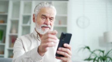 Evde oturma odasındaki kanepede oturan gri saçlı, akıllı telefonlu yaşlı bir adam. Olgun emekliler okur, mesajlar yazar, sohbet eder, online alışveriş yapar, telefonda başvuru kullanır. Kapat.