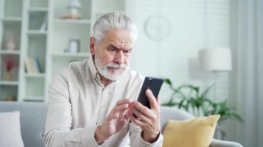 Evde oturma odasındaki kanepede otururken akıllı telefondaki yazılımla veya servisle ilgili sorunları olan sinirli yaşlı bir adam. Hayal kırıklığına uğramış olgun bir erkek kötü internet bağlantısından şikayetçi.
